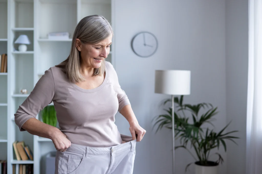 A smiling older woman holding oversized pants, demonstrating successful weight loss with the help of Tirzepatide.